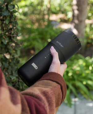 Elektresche Coffee Travel Mug schwaarz V2 - 355 ml, gëtt dobaussen an der Hand gehalen mat enger grénger, bléihender Hannergrond.